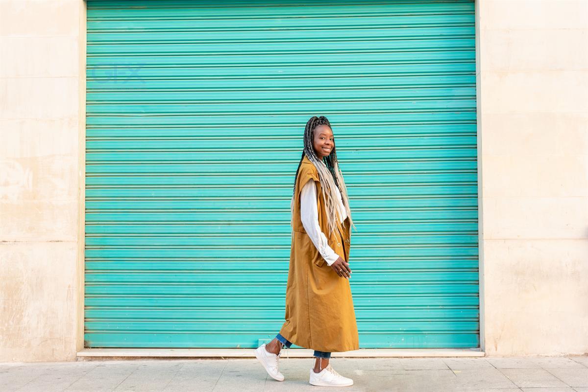 a young african american woman with braids walking on the street next to blue wall looking at camera