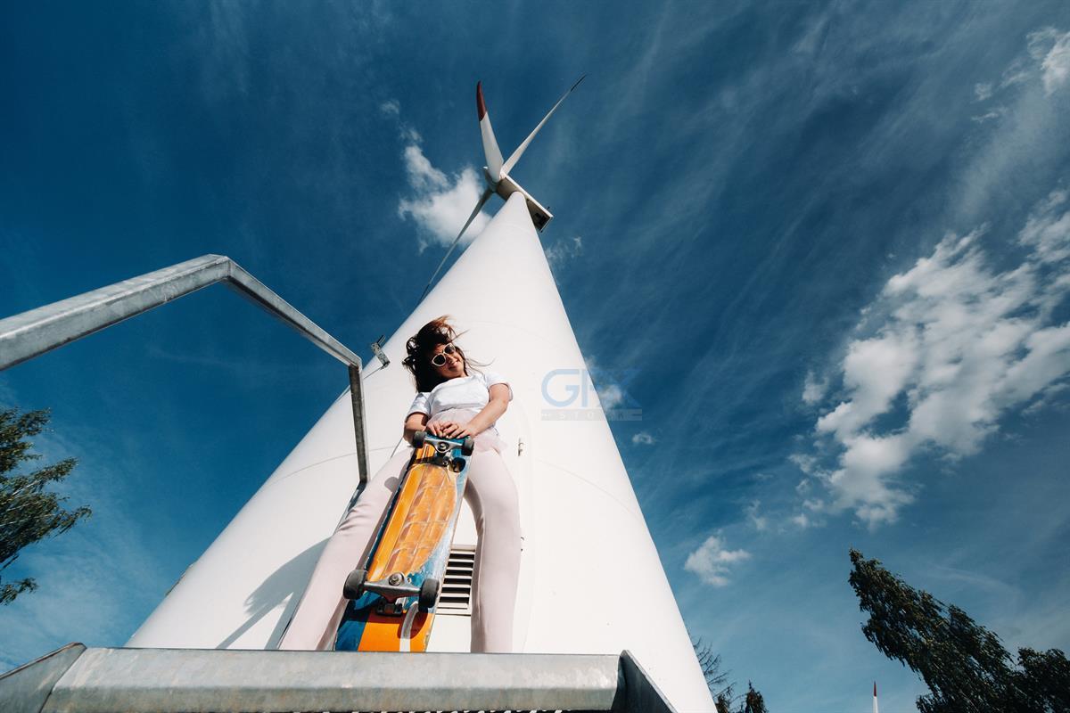 A girl in white clothes and glasses with a skate in her hands is photographed near large wind